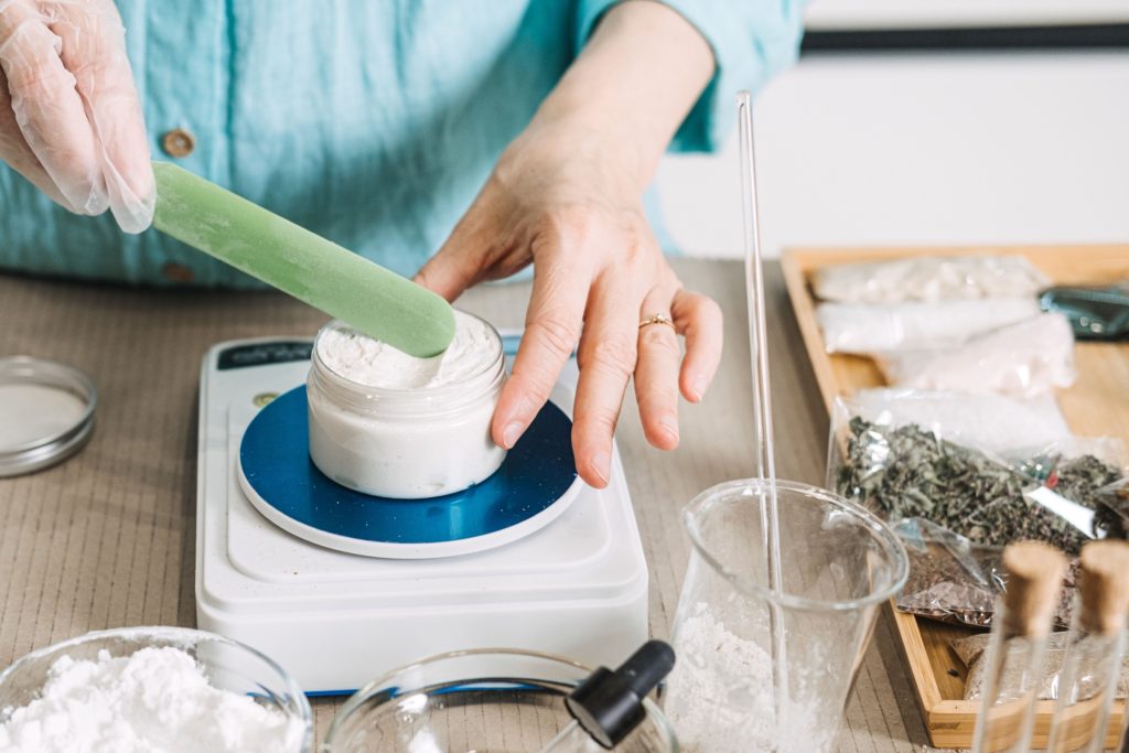 Hands use green spatula to mix white cream in jar on digital scale. Natural skincare formulation, chemical-free cosmetics, botanical ingredients, non-toxic beauty.
