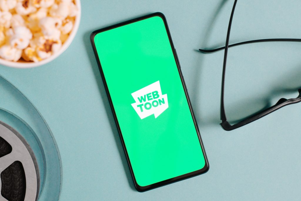 Webtoon South Korean platform mobile app on smartphone, popcorn cup, 3D glasses, film reel, top view.