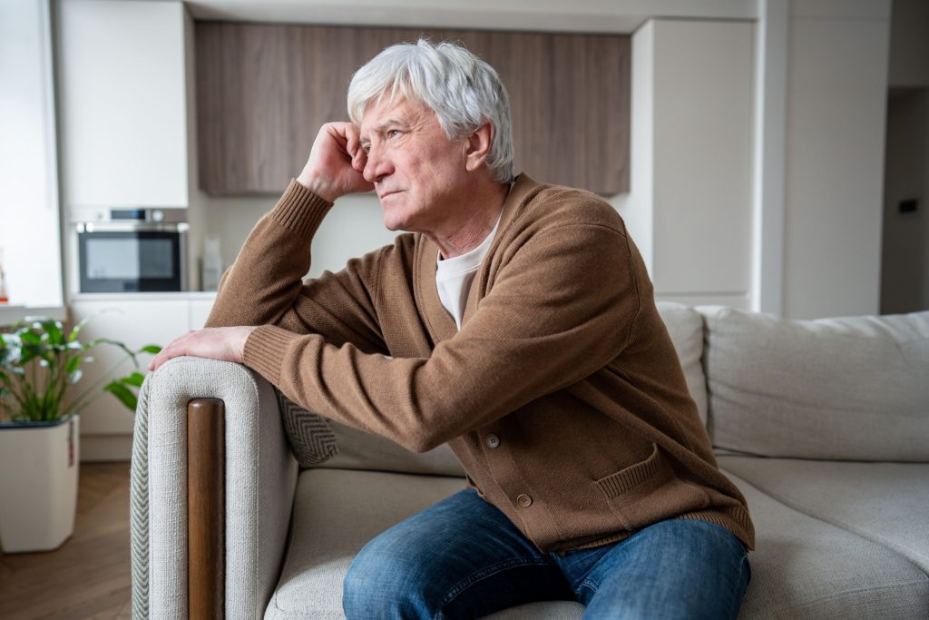 Aging senior man sits alone in empty living room staring vacantly, struggling with memory loss. Alzheimer confusion, chronic loneliness, cognitive decline, abandoned elder, neurological deterioration