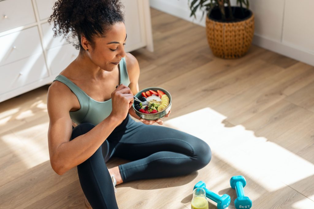 Shot of athletic woman eating a healthy fruit bowl while sitting on floor in the kitchen at home.